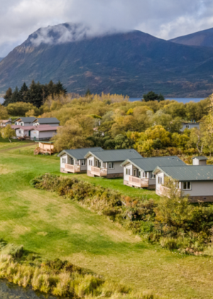 Small cabins with green roofs sit on a grassy hill at Kodiak Lodge, surrounded by trees, with a mountain in the background partially covered by clouds.
