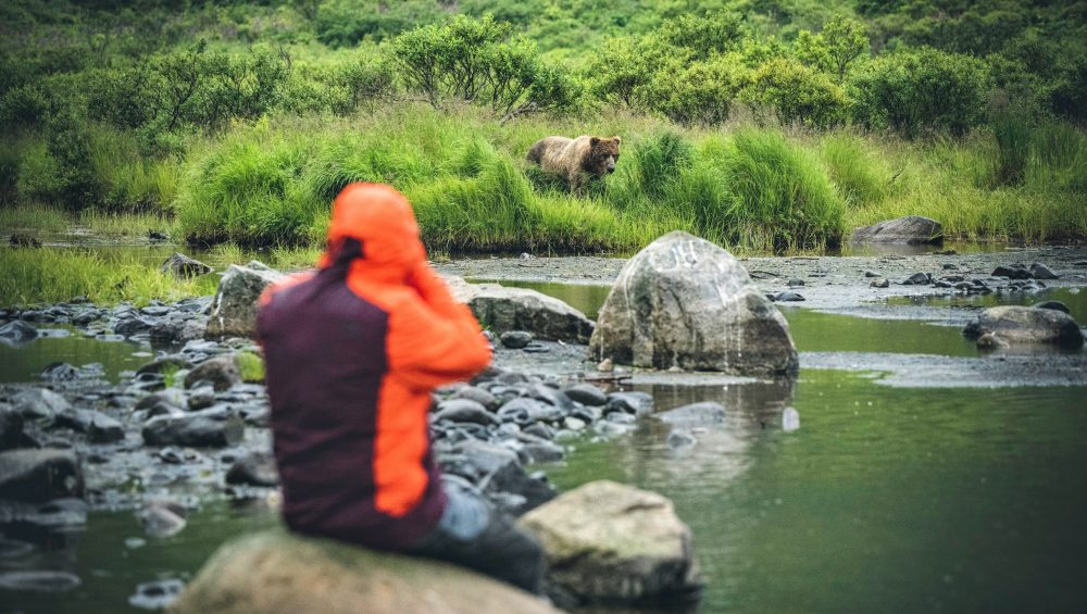 Dan M Lee in an orange jacket sits on rocks near a riverbank, photographing or observing a brown bear in the grass across the water, surrounded by green vegetation.