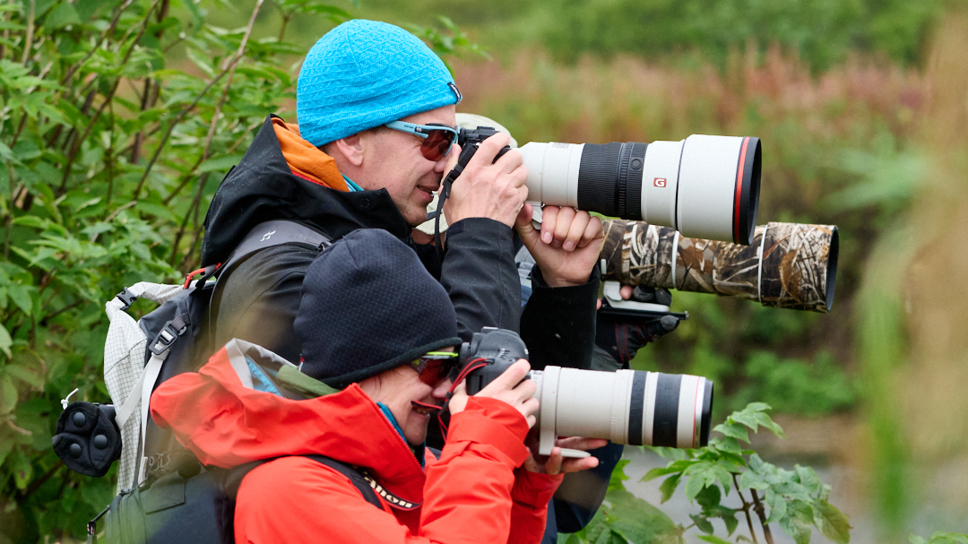Two people wearing colorful jackets and hats hold cameras with large telephoto lenses, aiming them forward while standing in a lush, green outdoor setting. Taken by Dan M Lee