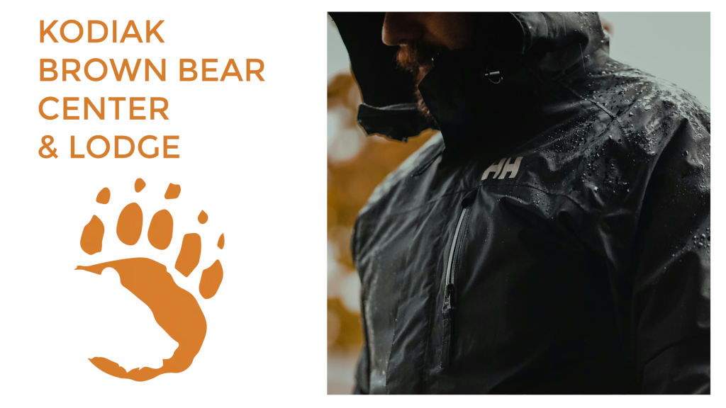 A person in a rain jacket stands outdoors next to text reading Kodiak Brown Bear Center Lodge with an orange bear paw print graphic.