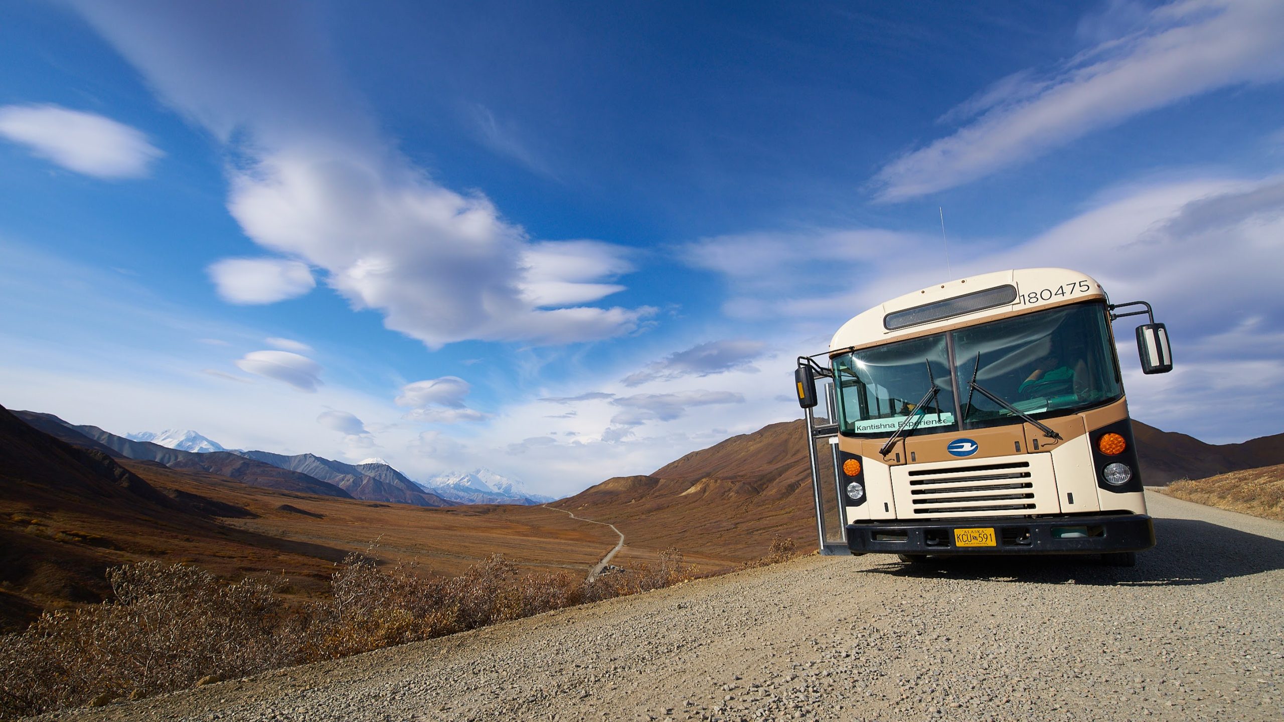 A white bus travels along a gravel road through a vast, open landscape with brown hills and distant snow-capped mountains under a blue sky with scattered clouds.